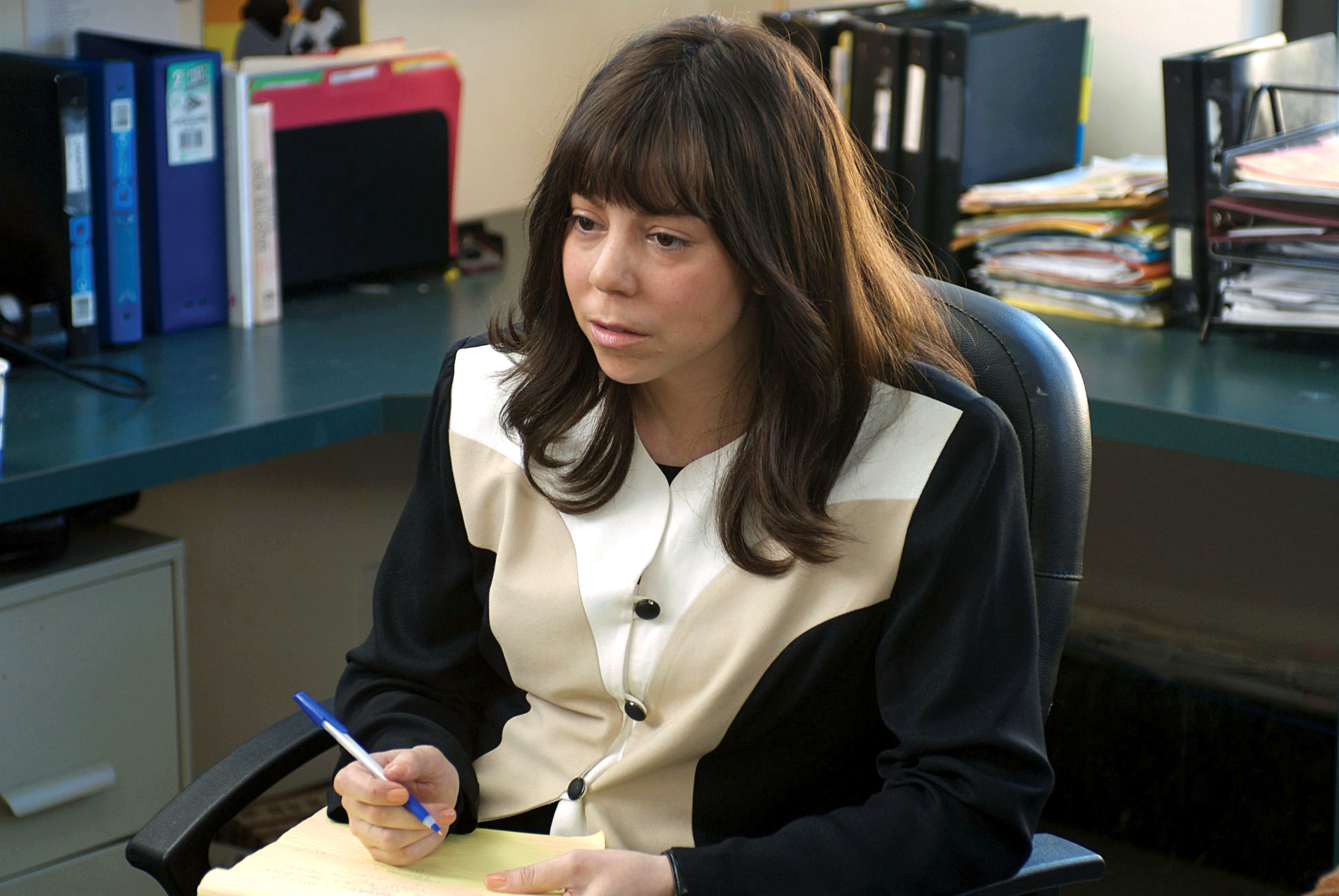 America Ferrera sits at a desk, holding a pen and notepad, dressed in a buttoned blouse with contrasting fabric design