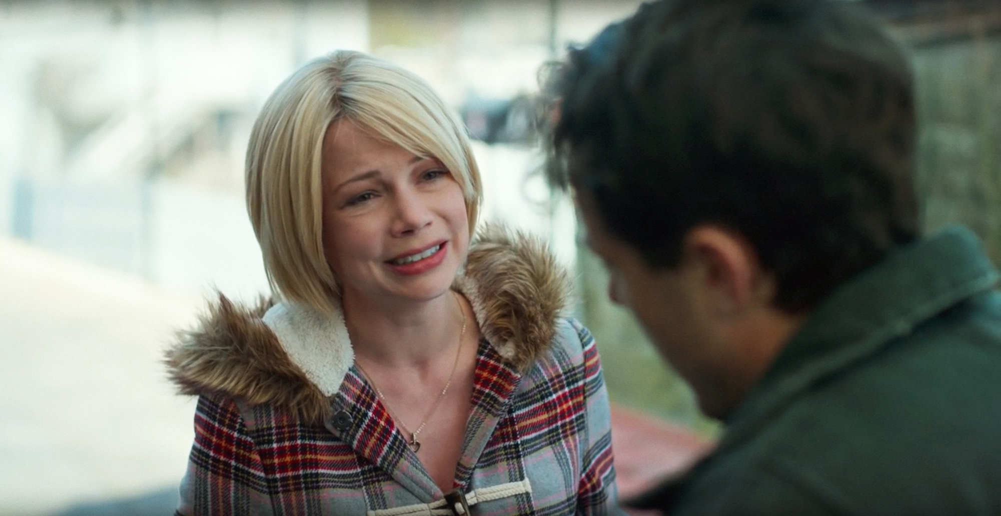 Michelle Williams in a plaid coat with a fur-trimmed hood talks to Casey Affleck, who has his back to the camera. Scene from TV/movie
