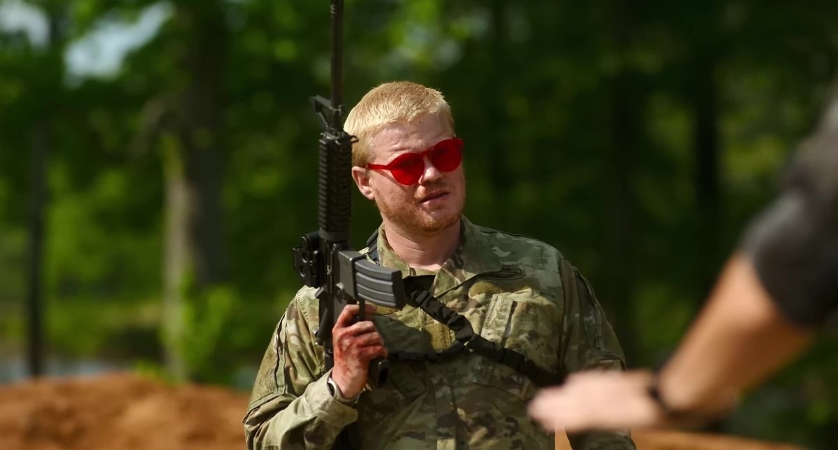 A man in military uniform holds a rifle and wears red-tinted sunglasses, standing outdoors with trees in the background