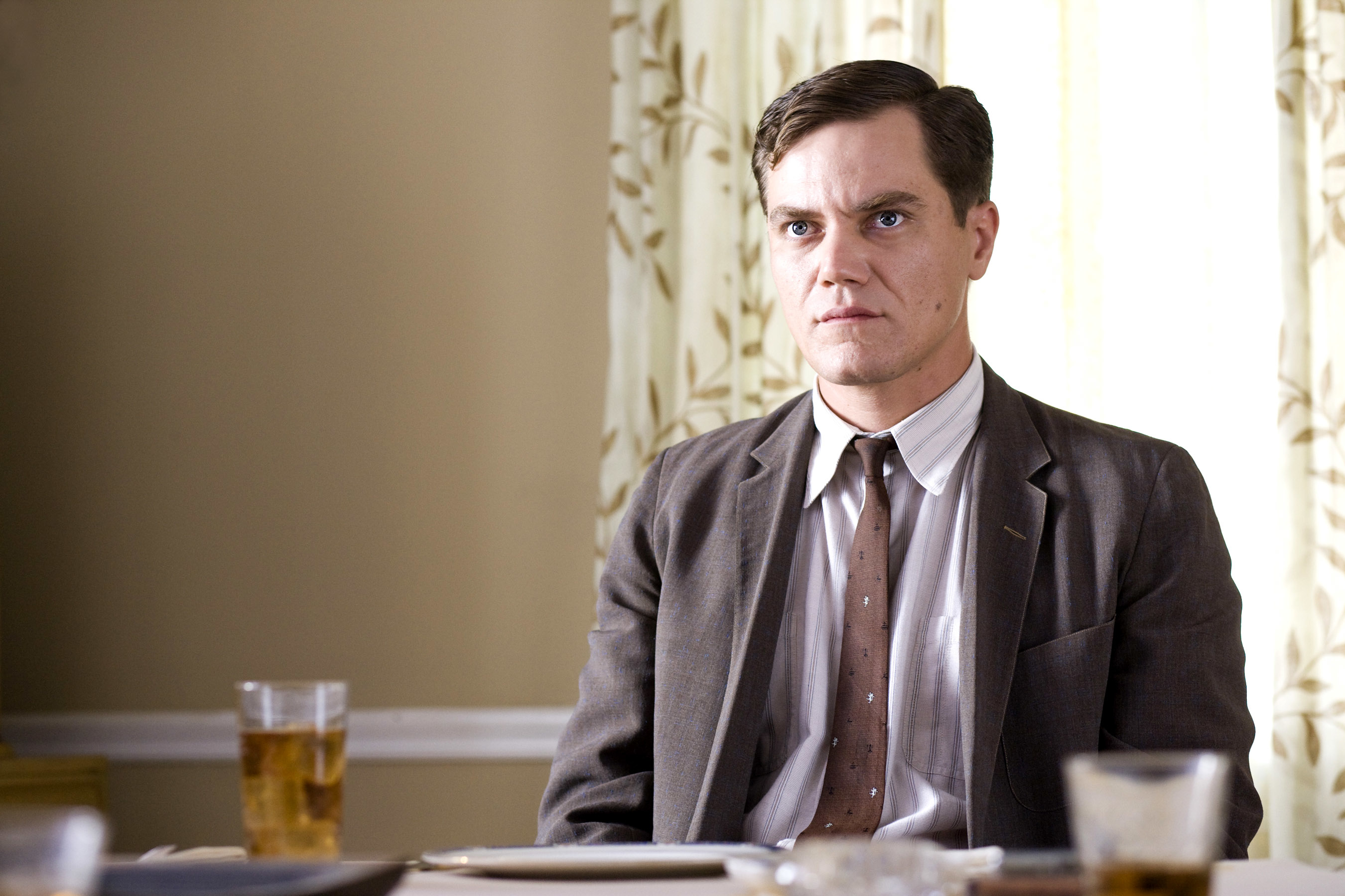 Man in a suit and tie sits at a dining table with a glass of tea, appearing serious. There are curtains with a leaf pattern in the background