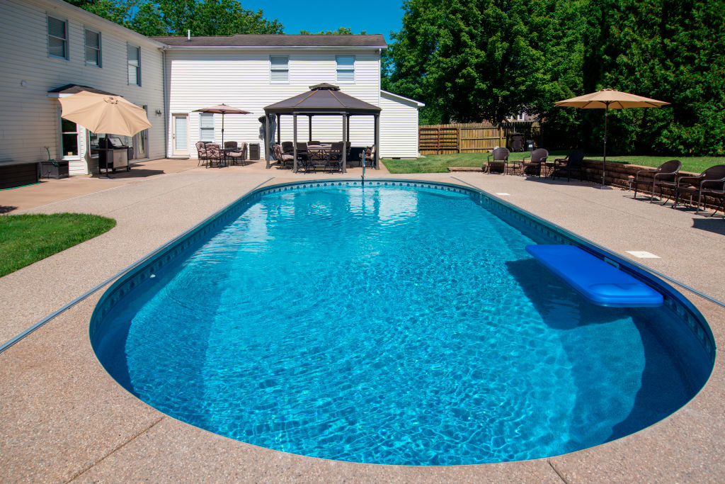 Backyard with an in-ground swimming pool, surrounded by lounge chairs, umbrellas, and a gazebo. A large white house is in the background