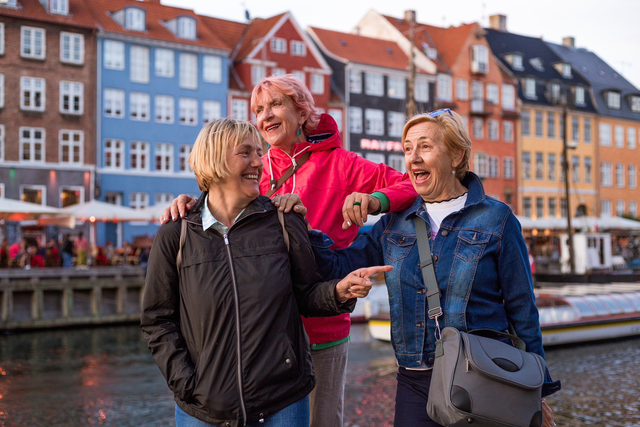 Three women laughing and enjoying their time by a waterfront with colorful buildings in the background in Copenhagen, Denmark