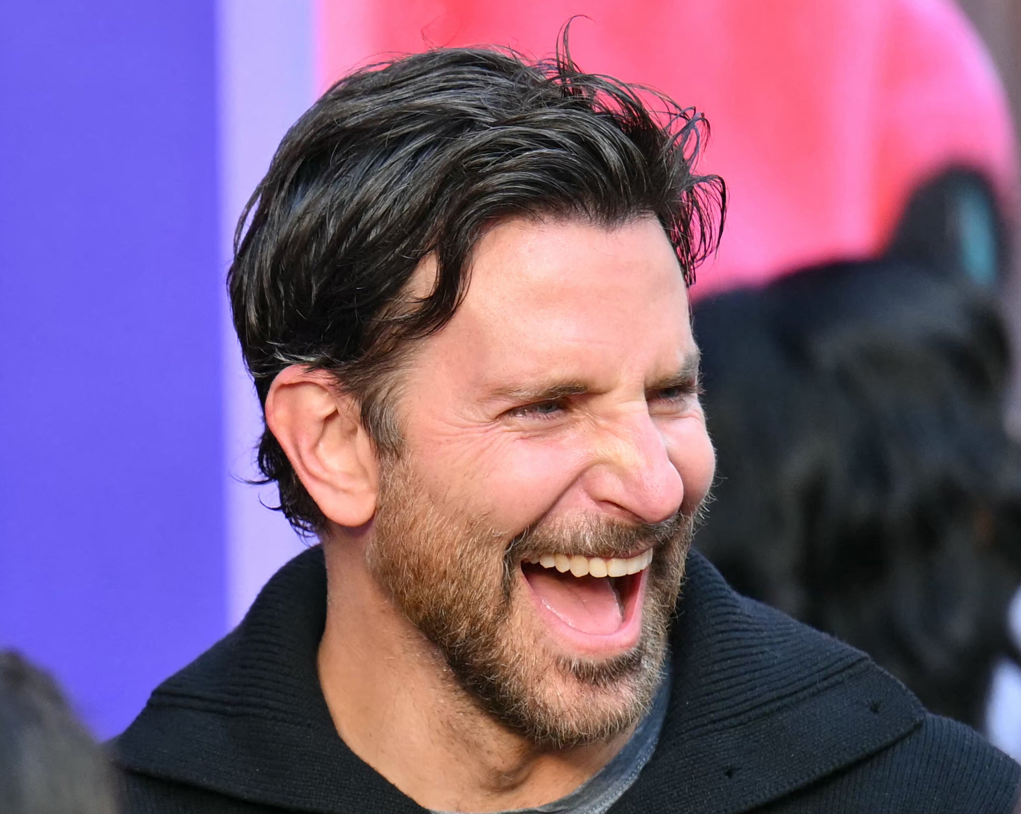Bradley Cooper laughs with a bearded man at a public event, with another person in glasses and curly hair smiling in the background