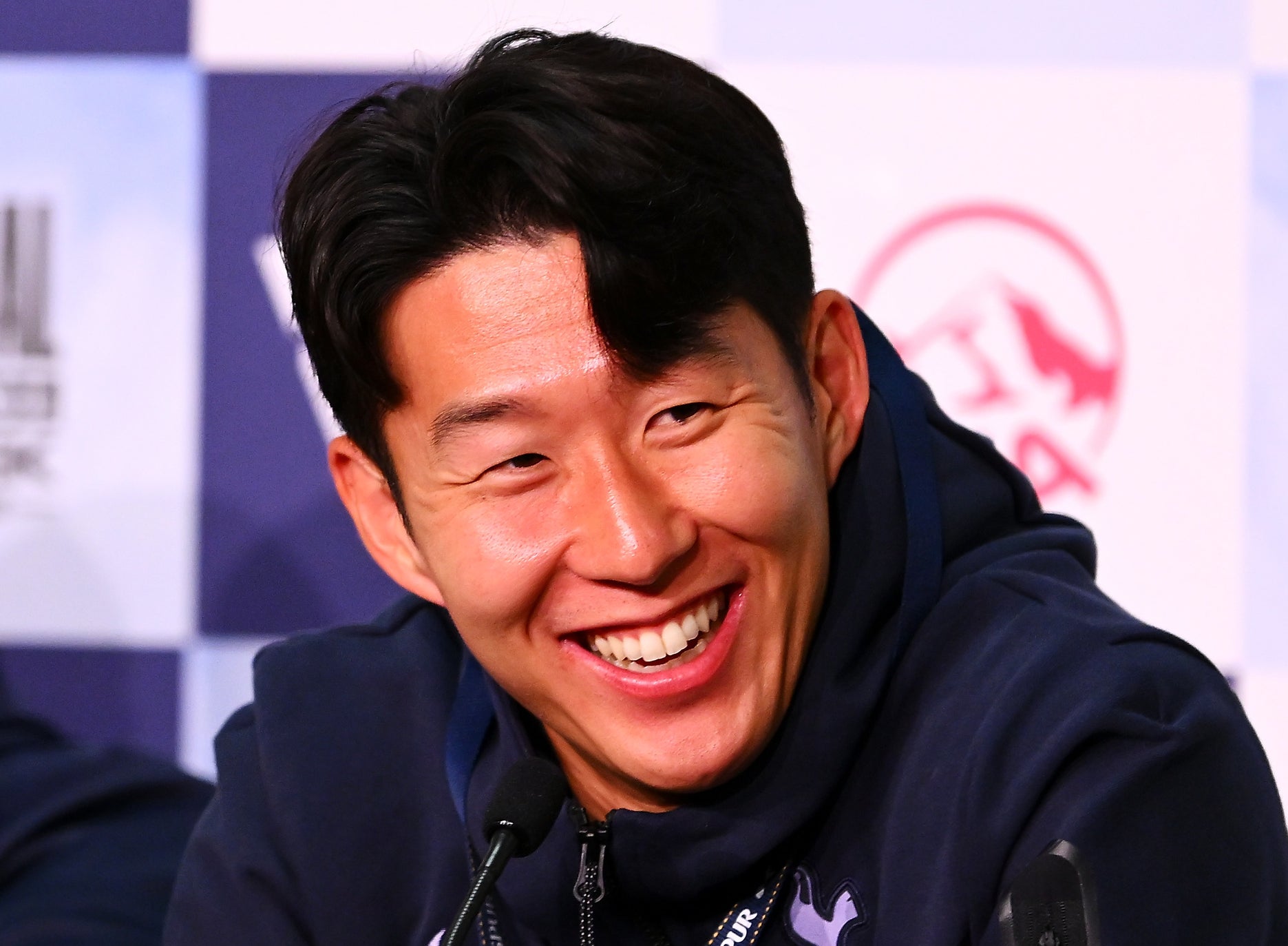 Son Heung-min and coach Ange Postecoglou, in casual sportswear, speak and smile at a press conference with various microphones on the table