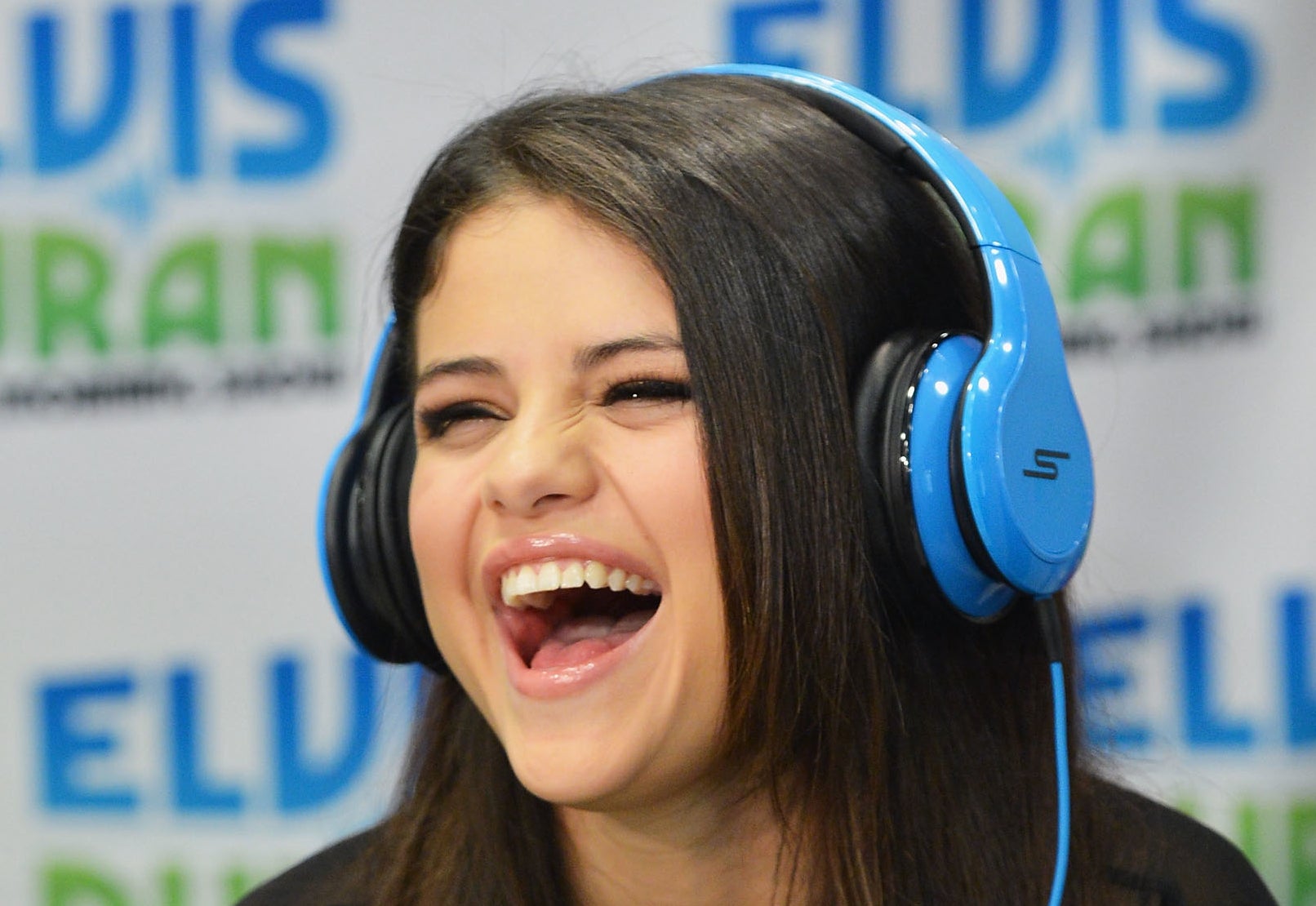 Selena Gomez laughs while wearing blue headphones and speaking into a microphone during an appearance on "Elvis Duran and the Morning Show."