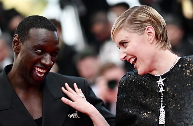 Omar Sy and Greta Gerwig share a joyful moment on the red carpet; Omar in a black suit, Greta in a glittering black dress with a cape and statement jewelry