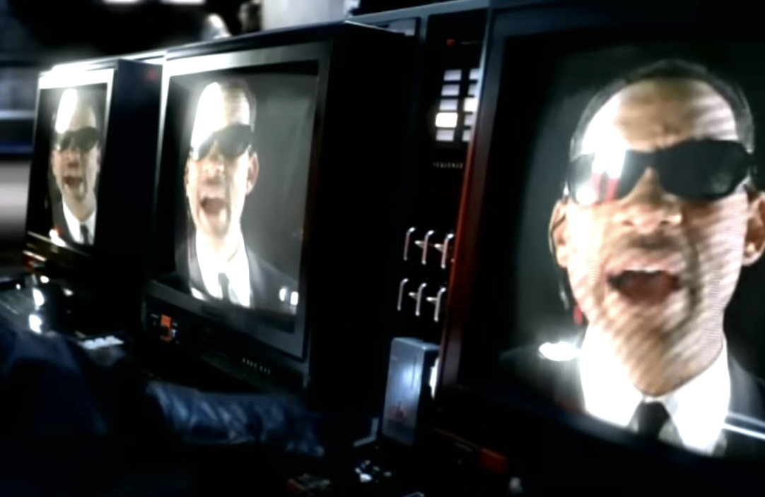 Will Smith in sunglasses appears multiple times on screens in a control room