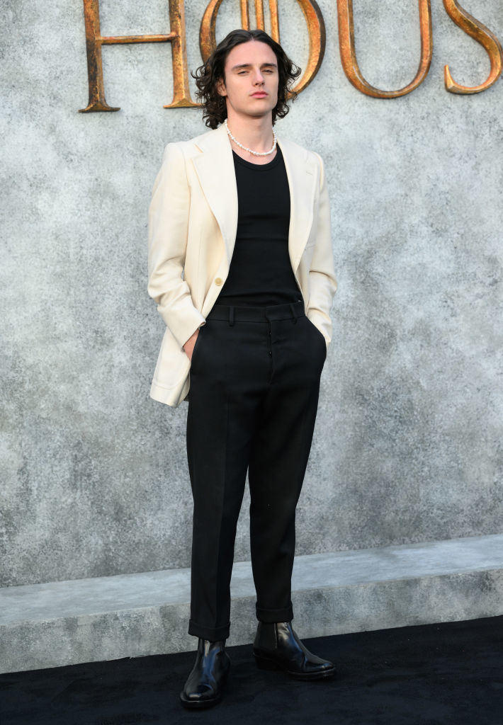 Harry Collett stands on a red carpet wearing a cream blazer, black shirt, black trousers, and black shoes