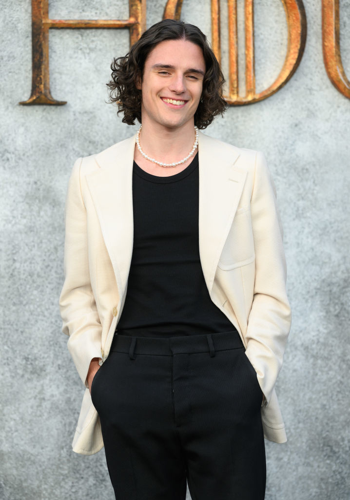 Harry Collett with shoulder-length curly hair wearing a black shirt, cream-colored blazer, black pants, and a pearl necklace. Background is blurred