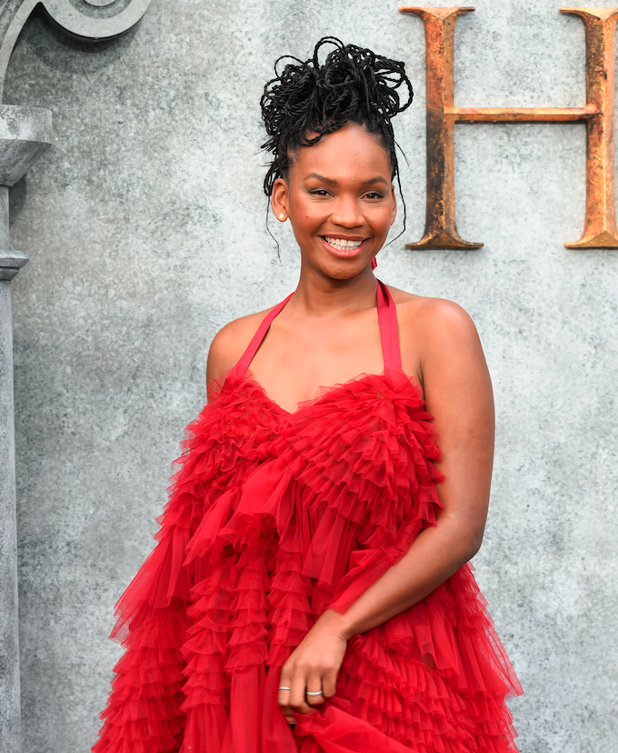 Bethany Antonia in an elegant red, layered ruffle dress, smiling in front of a stone wall with a decorative "H" behind her