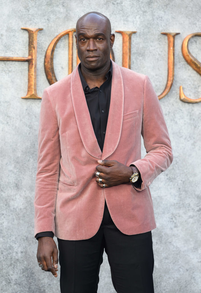 Steve Toussaint in a velvet blazer and dark shirt stands in front of a backdrop with large, gold text partially visible