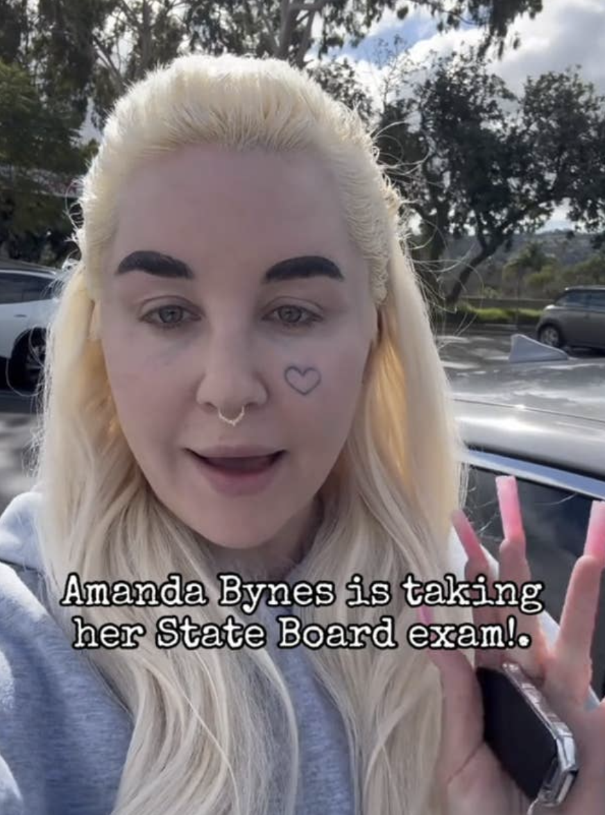 Amanda Bynes outdoors with long hair, heart face tattoo, and nose ring, holding a pen. Text: "Amanda Bynes is taking her State Board exam!"