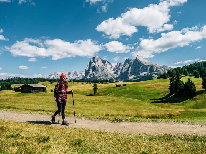 A person with hiking gear walks along a dirt path in a mountainous countryside with green fields and a few scattered buildings in the background