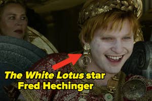 Fred Hechinger, dressed in Roman-style attire with a large earring, smiles in a scene from "The White Lotus." A red arrow points to him