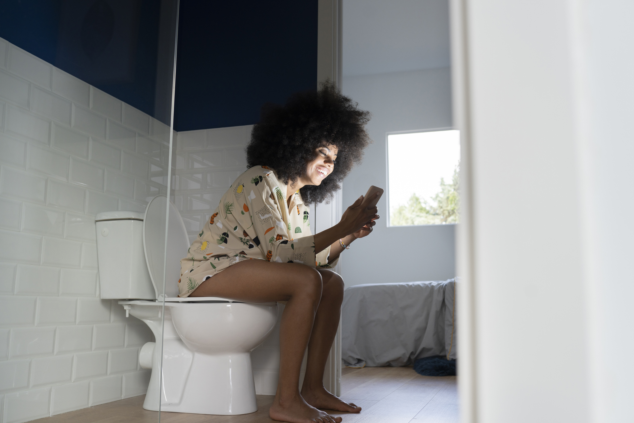 Person sits on a toilet, smiling while looking at a phone. They are wearing patterned loungewear. The bathroom has a window providing natural light