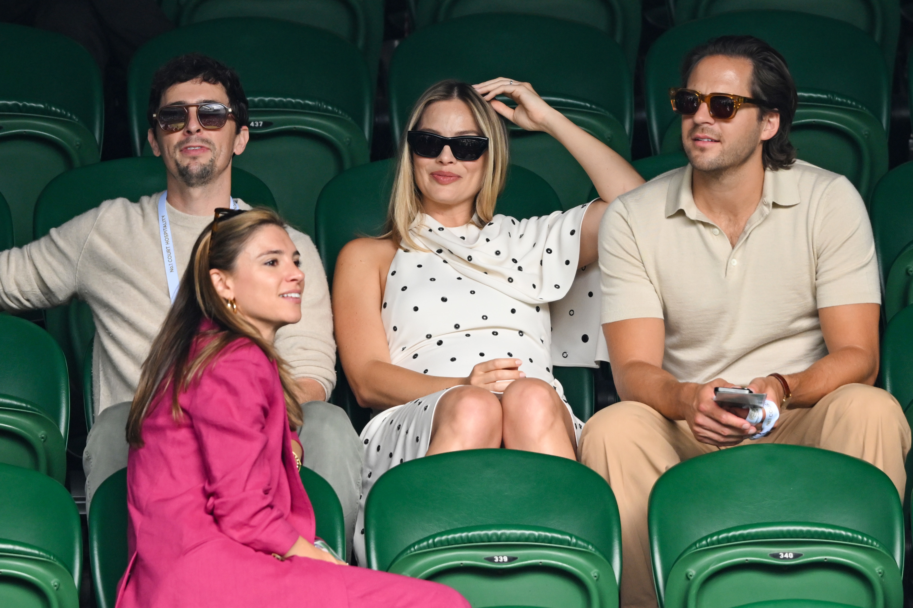 Margot Robbie, Tom Ackerley, and two friends sitting in green stadium seats, dressed casually with sunglasses