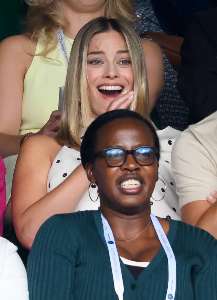Margot Robbie smiles with hands clasped in front of her face. A person with glasses is seated in front, smiling with an open mouth