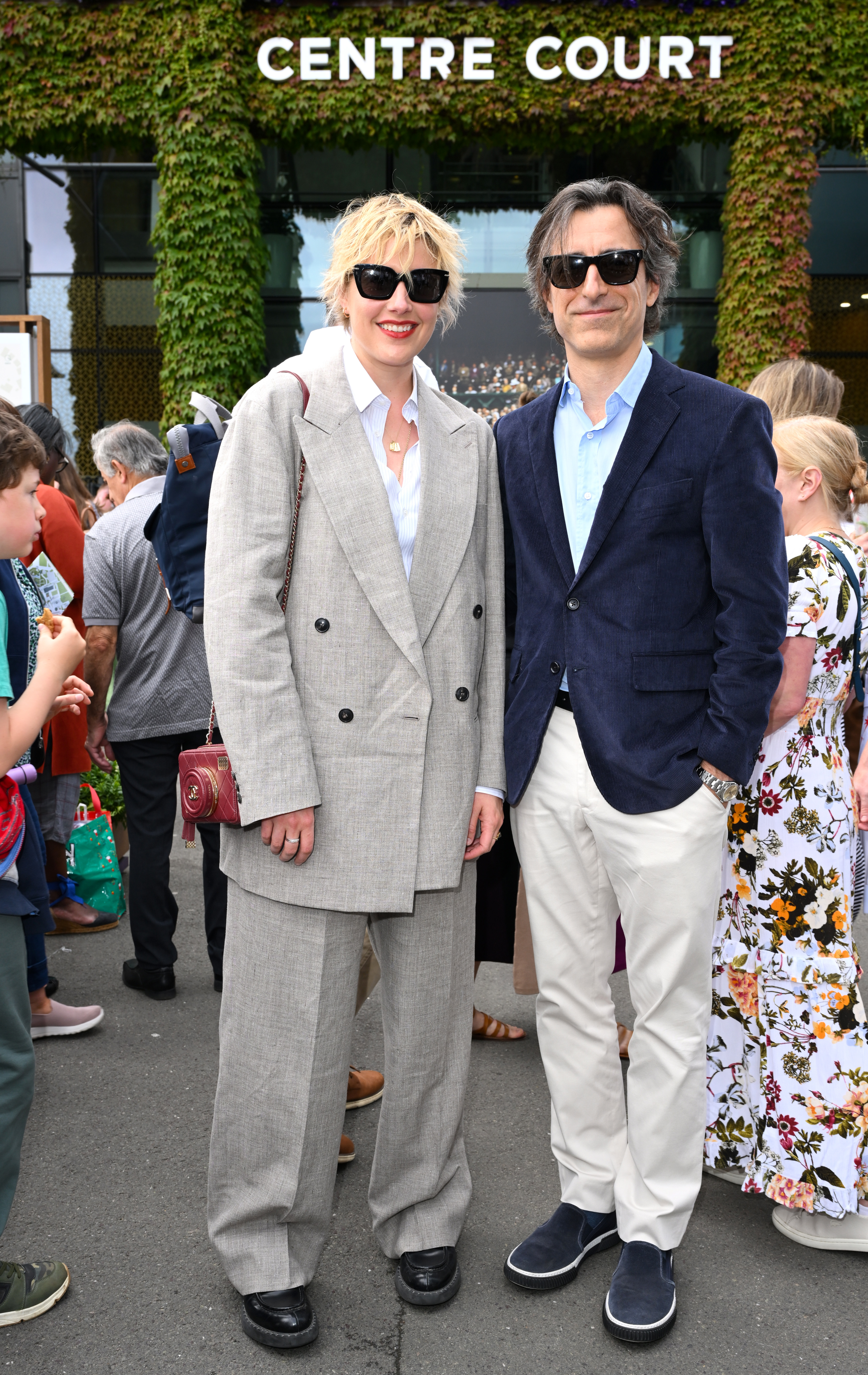 Greta Gerwig in a oversized suit and black shoes, and Noah Baumbach in a blue blazer with light pants and sneakers, standing outside Centre Court