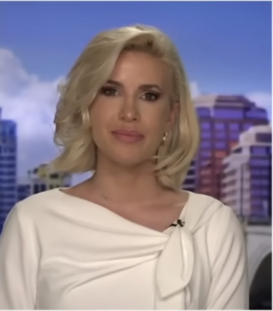 A woman with short blonde hair, possibly a news anchor, is wearing a white, elegant top with a bow near the shoulder. The background shows a cityscape