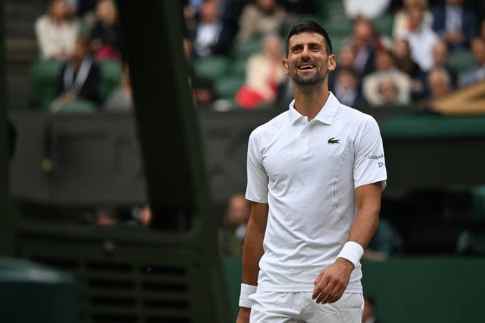 Novak Djokovic on a tennis court, smiling and wearing a white polo shirt and shorts. He appears to be at a professional tennis event