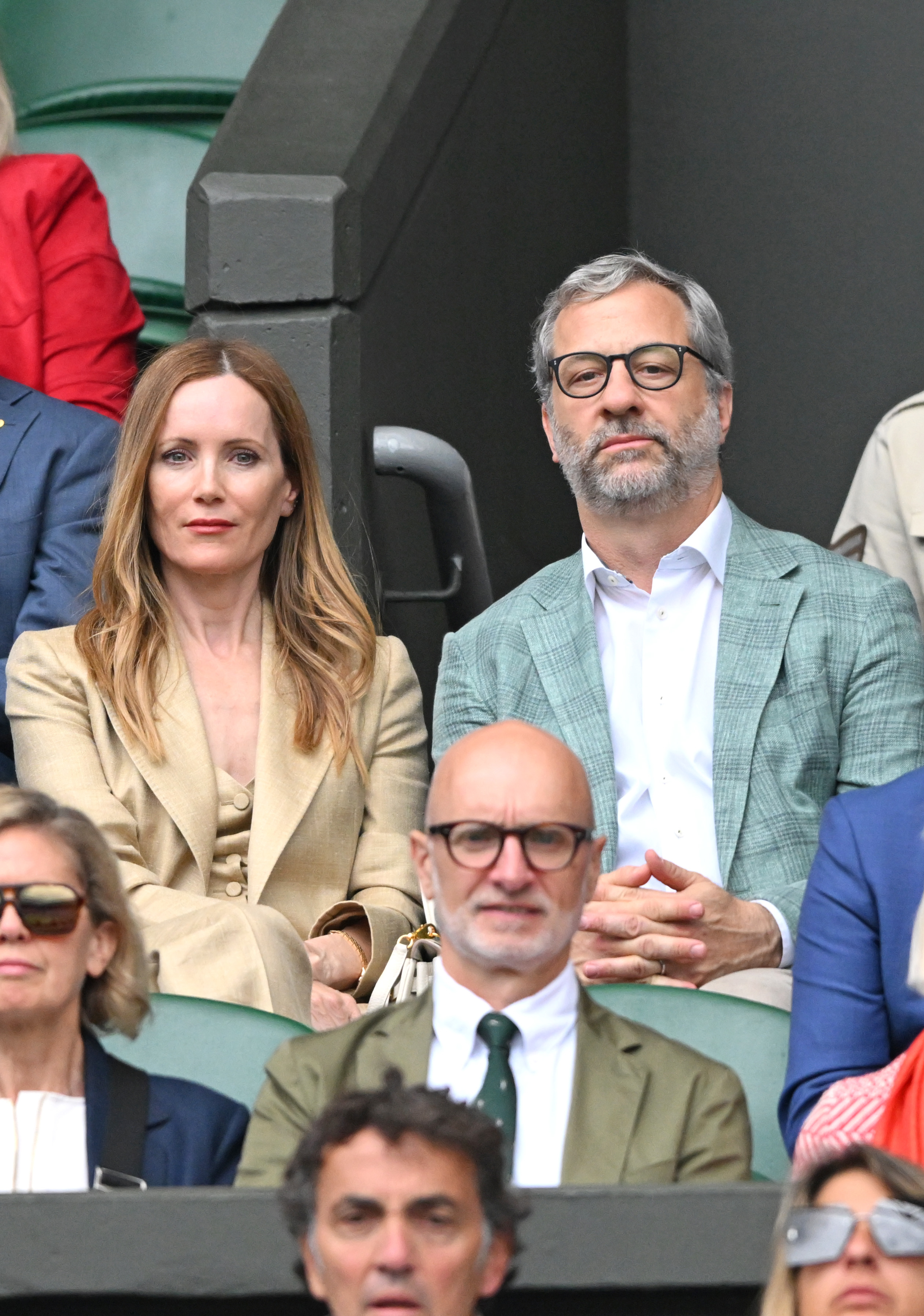 Leslie Mann in a tan outfit and Judd Apatow in a light green blazer sit together at a public event, a crowd is in the background