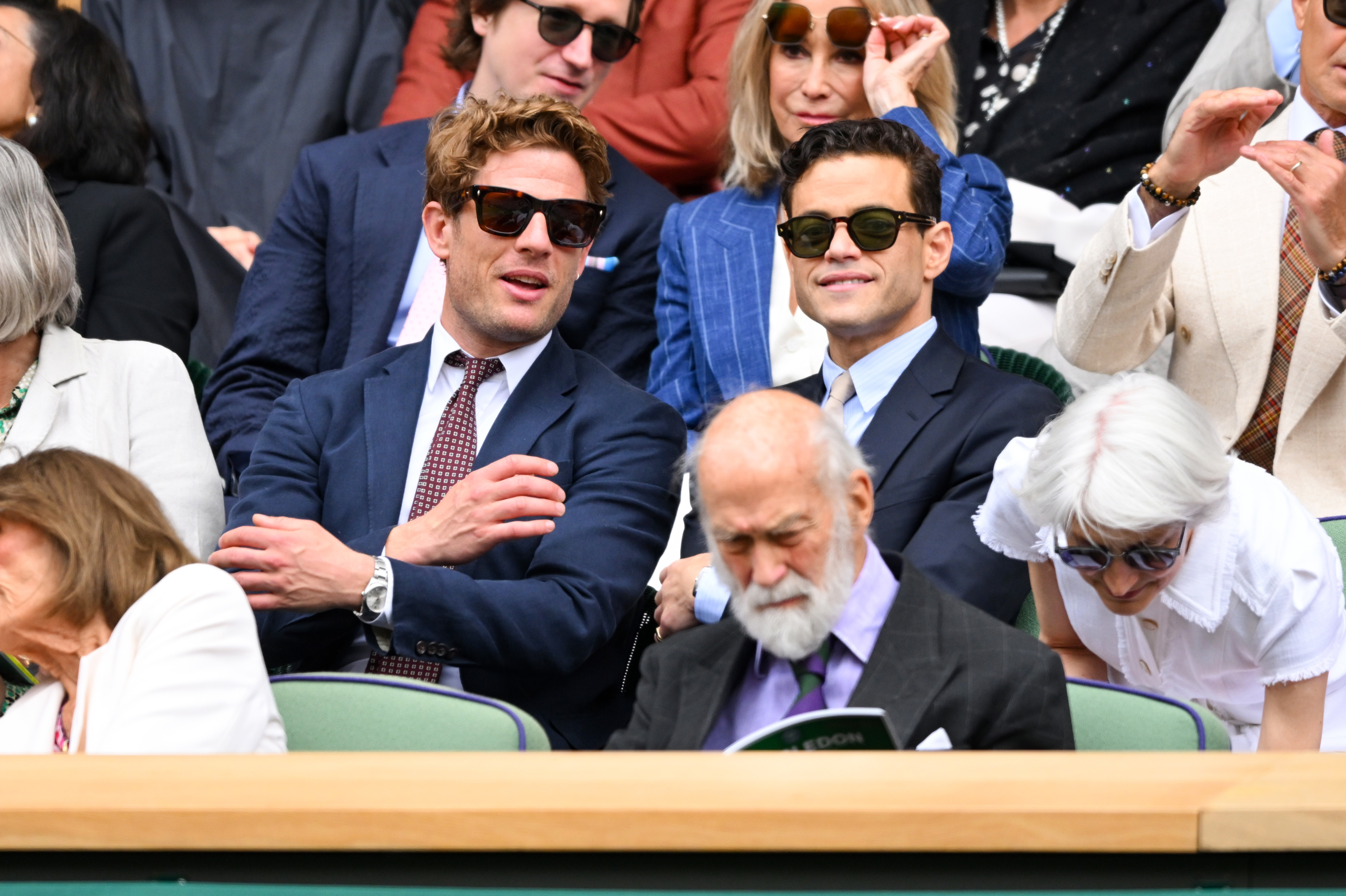 James Norton and Rami Malek, in suits and sunglasses, sit in the audience at Wimbledon. People around them are seated and looking at the event