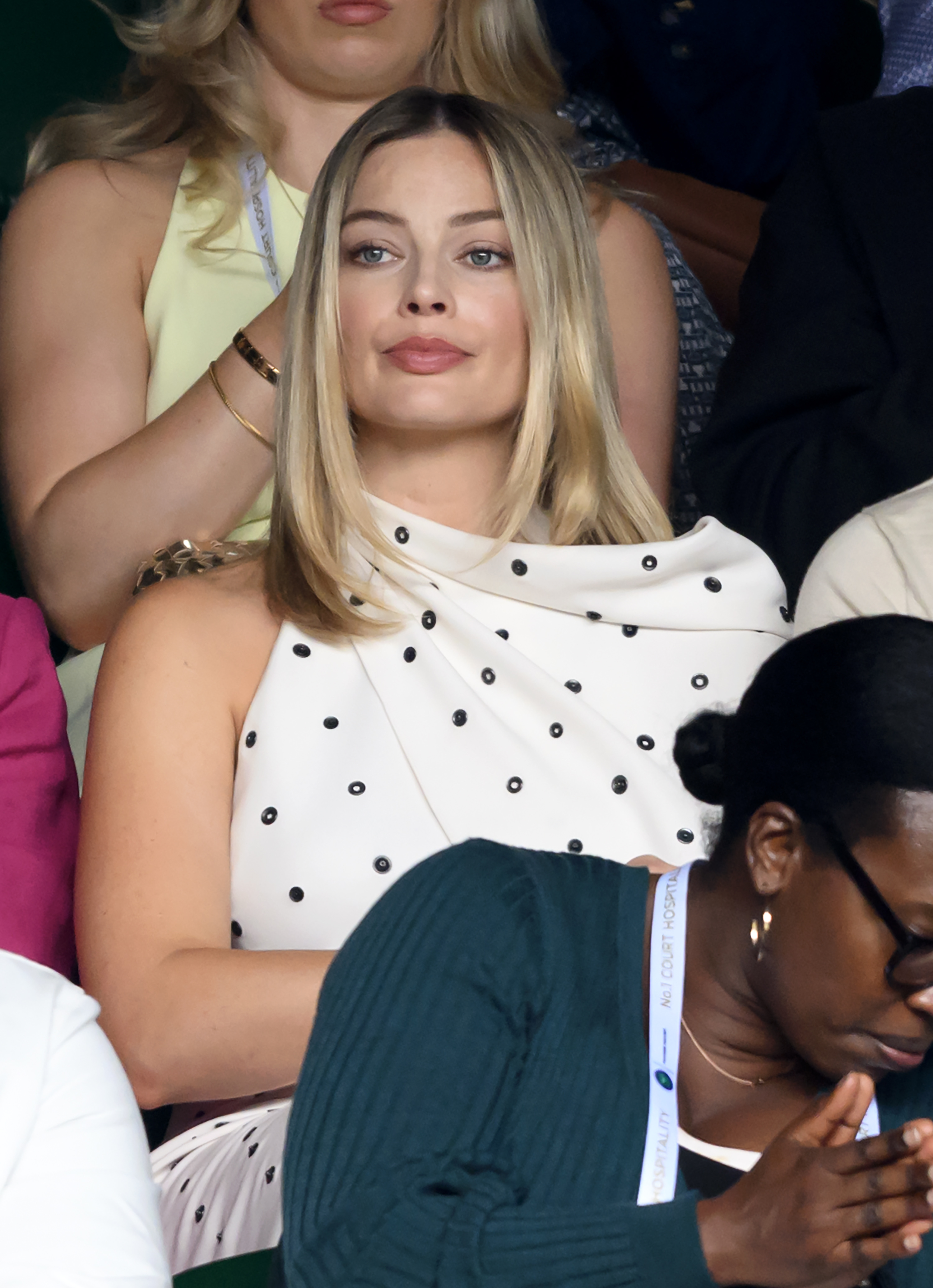 Margot Robbie sitting in the audience, wearing a stylish one-shoulder white dress with black polka dots. Other attendees are around her