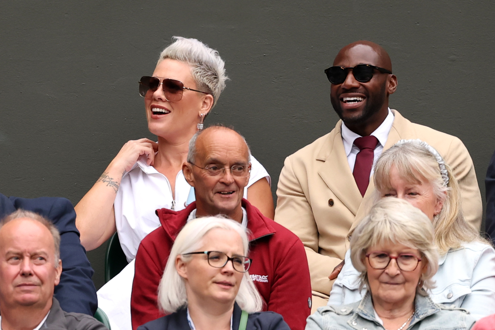 Pink and former athlete Derek Chisora are seated among others at an event. Pink is wearing a white outfit and sunglasses; Derek is in a beige suit with sunglasses