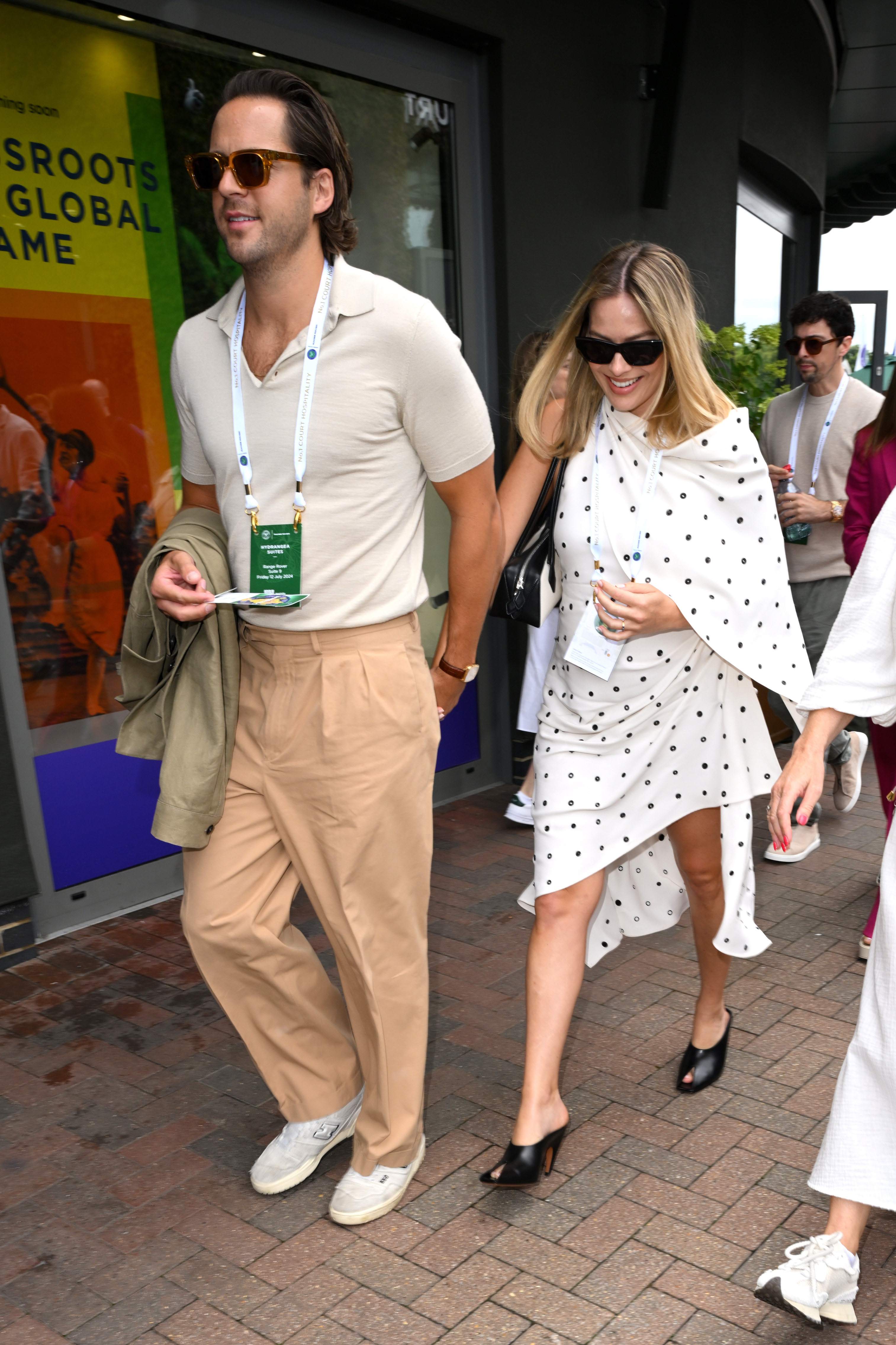 A man and a woman wearing sunglasses and badges walk outdoors. The man is in a casual shirt and pants, and the woman is in a polka-dotted dress with black shoes