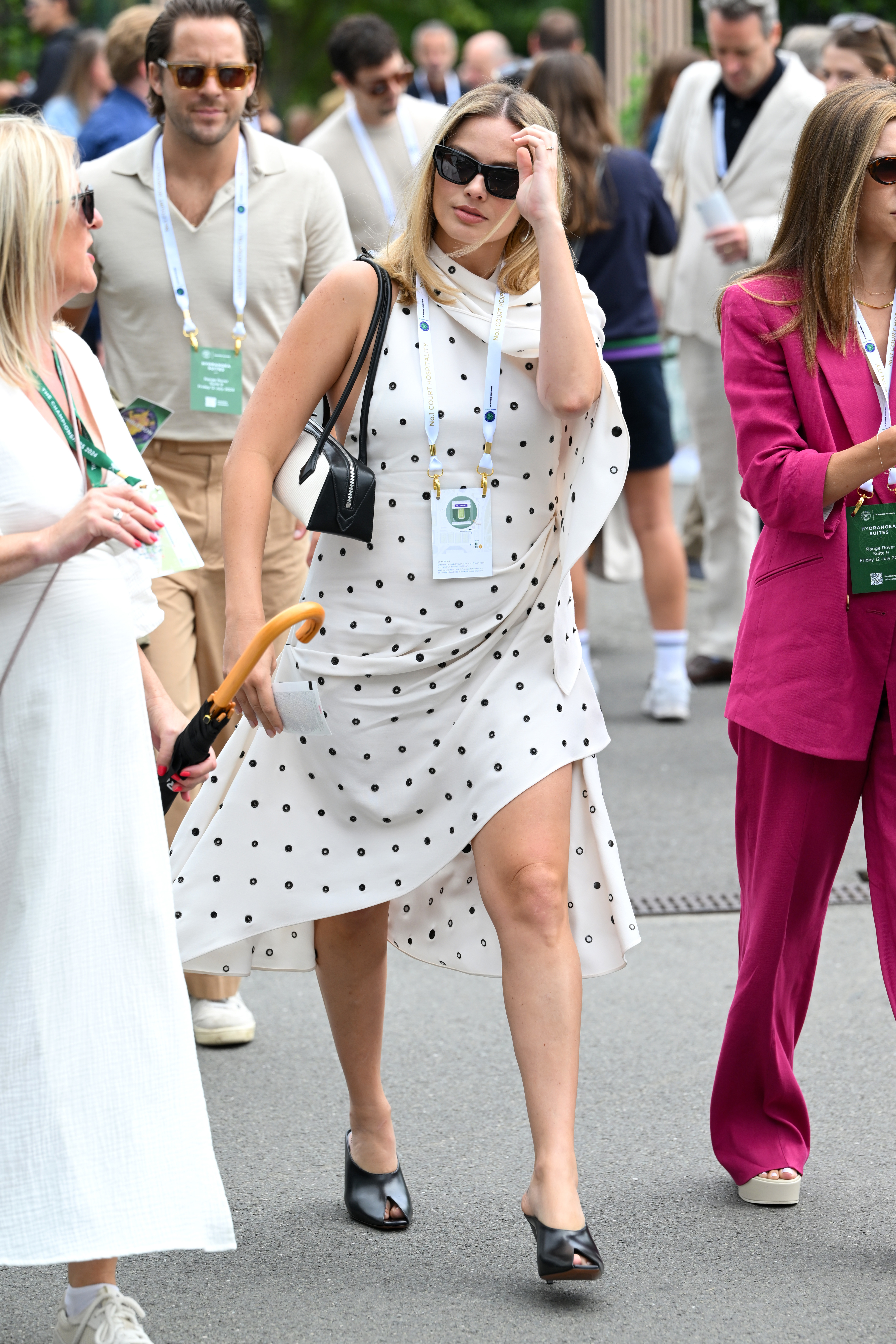 Margot Robbie on a sidewalk, wearing a polka-dot dress and black heels, carrying a black handbag, accompanied by multiple people