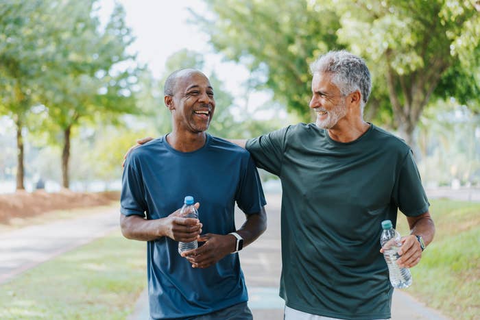 Two men, one bald and one with gray hair, wearing casual athletic wear, walking outdoors with water bottles, smiling and engaging in conversation