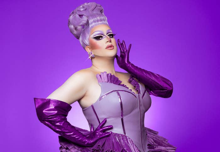 Drag queen in extravagant costume posing with one hand on her face and the other on her waist against a solid background