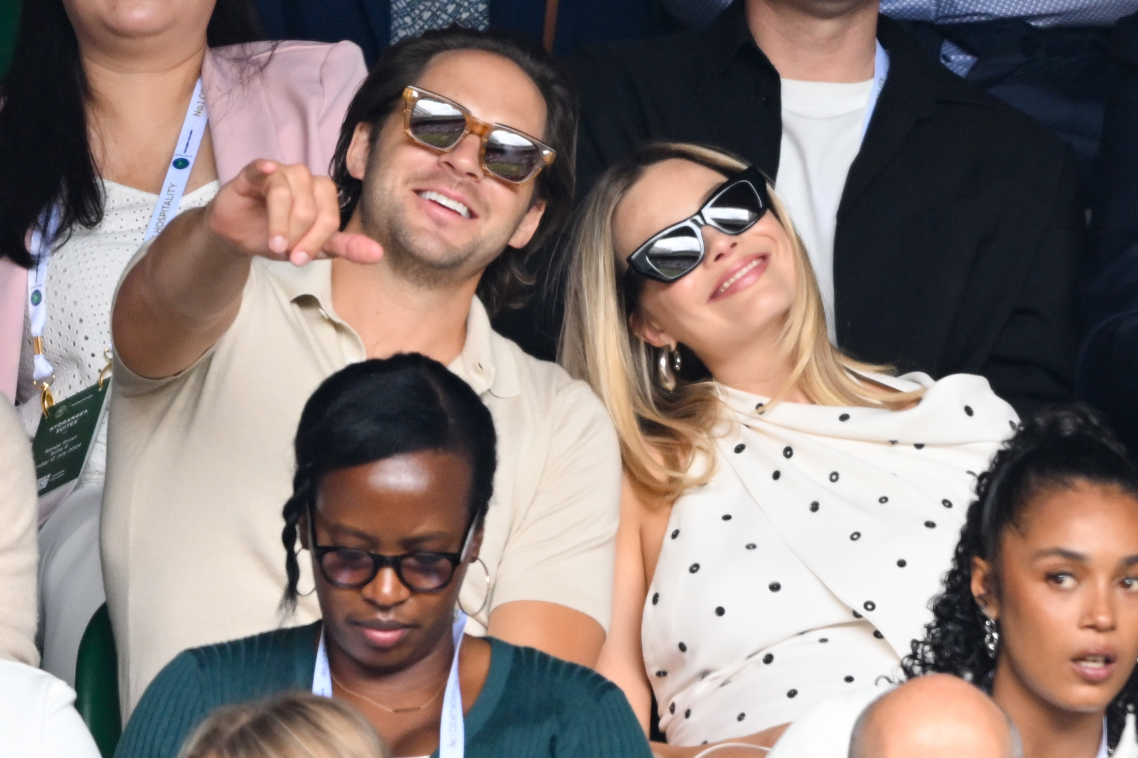 A group of people sitting in an audience. Two people, dressed casually with sunglasses, smile and point at something. Other individuals around them are watching attentively