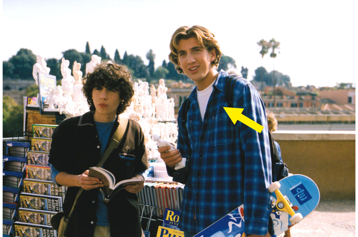 Two young individuals, one with curly hair holding a book, and another with wavy hair holding a skateboard and a blue plaid shirt, appear in an outdoor setting