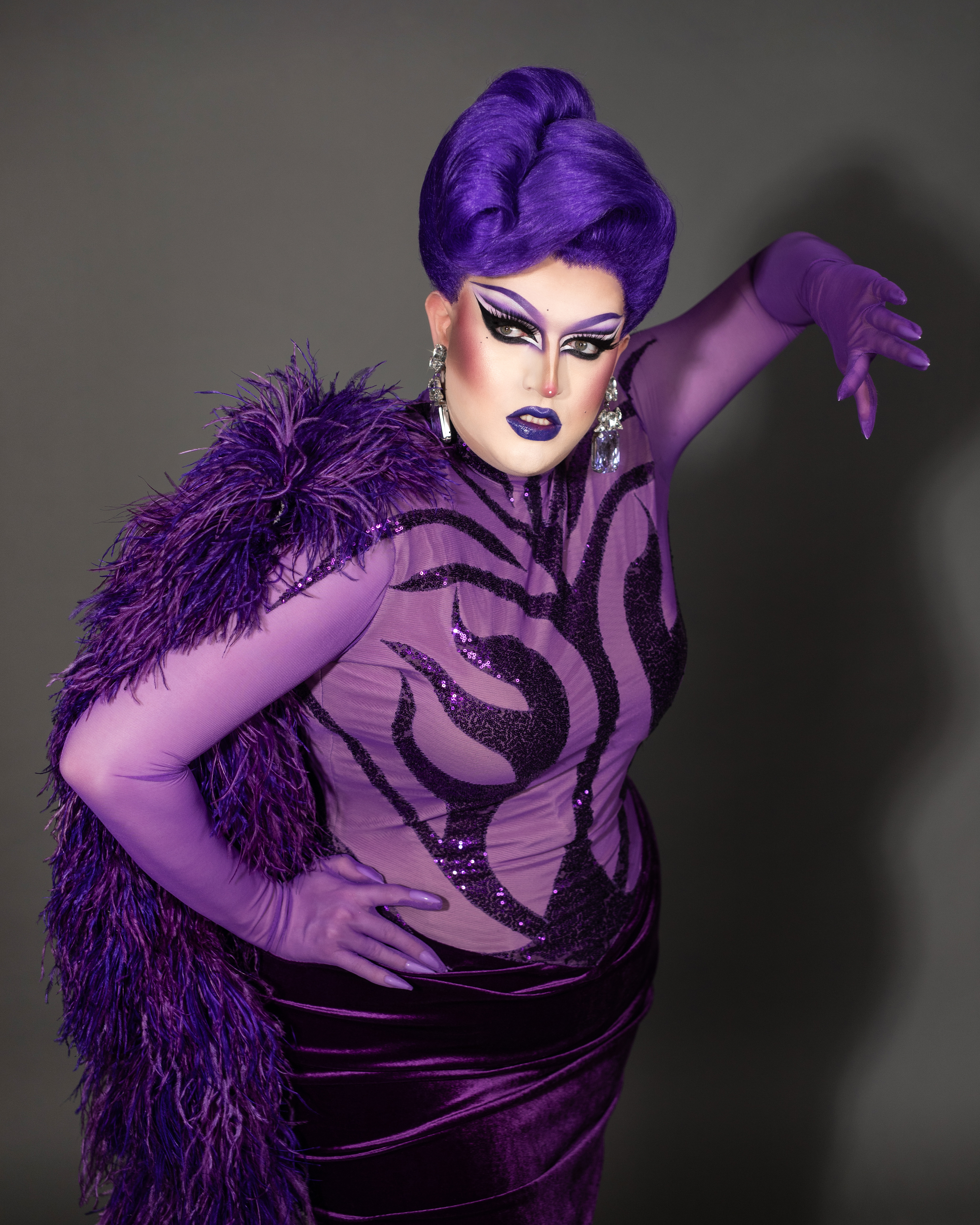 Drag queen in stylized purple velvet and sequined gown strikes a dramatic pose with intense makeup and a feather boa