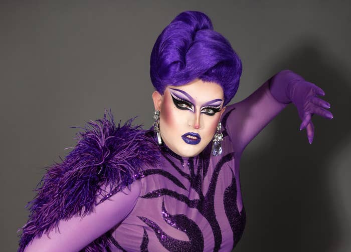 Drag queen in stylized purple velvet and sequined gown strikes a dramatic pose with intense makeup and a feather boa