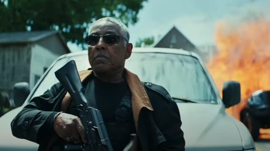 Giancarlo Esposito stands in front of a burning vehicle, holding a rifle, wearing sunglasses, a black jacket, and a brown scarf