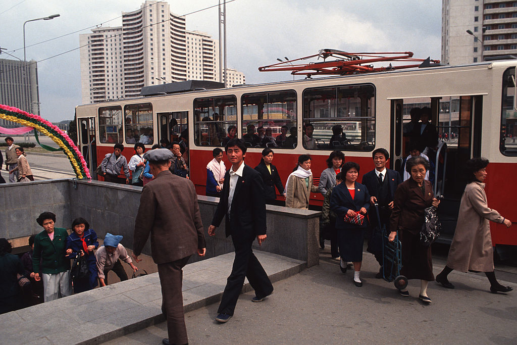 People disembark a tram and others enter a subway station in a bustling urban area, surrounded by tall buildings