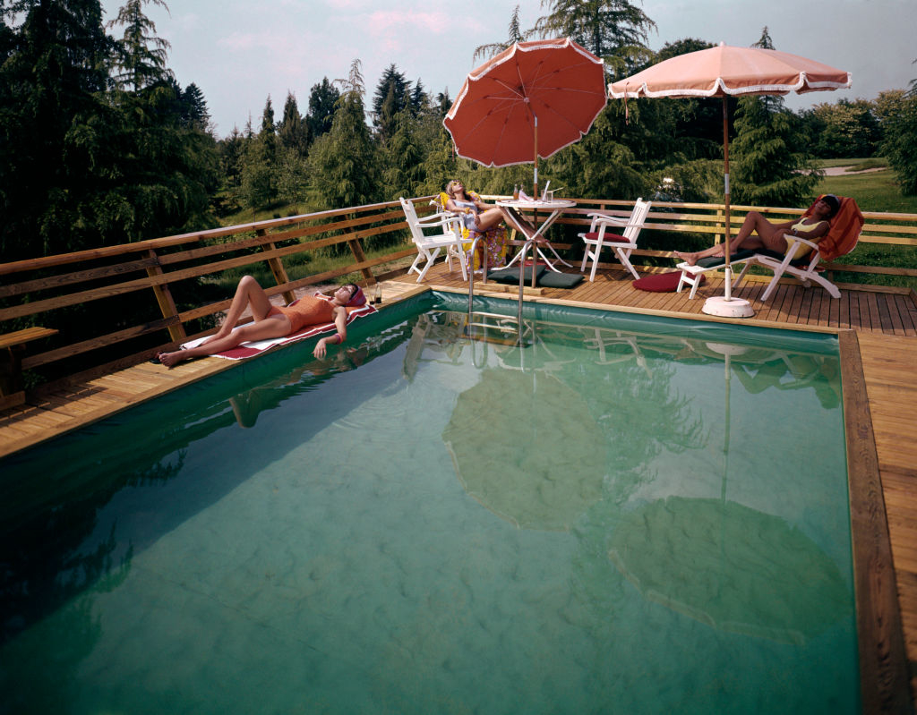 Three people relaxing poolside, one seated at a table under an umbrella, one lounging in a chair, and one lying on the pool edge. Trees surround the pool area