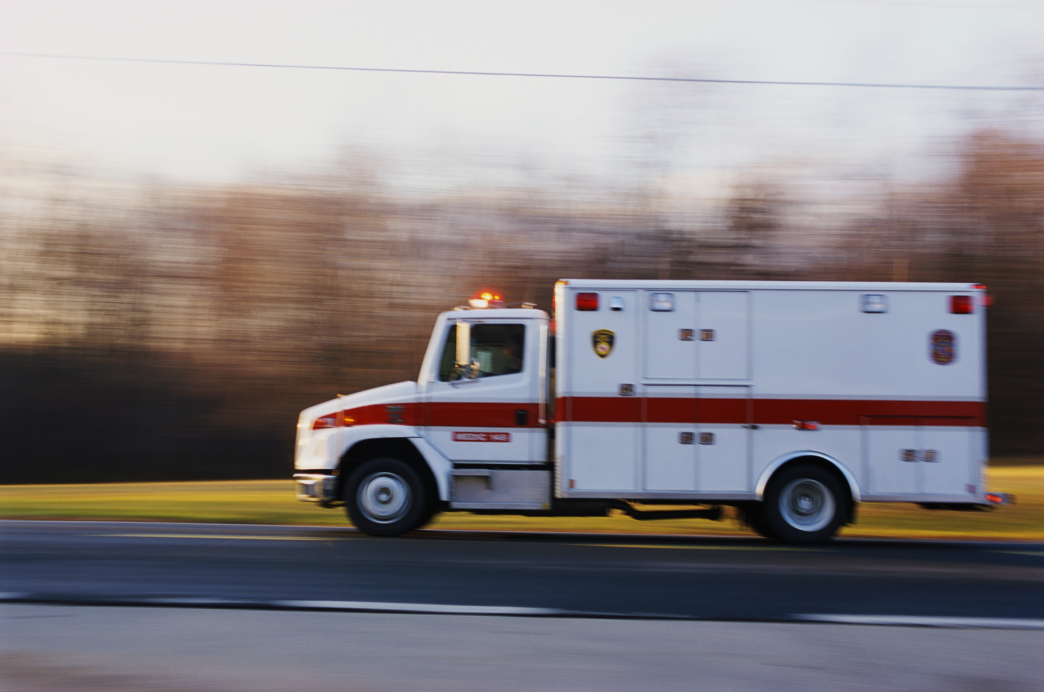 An ambulance with emergency lights on is driving quickly on a road, with a blurred background indicating speed