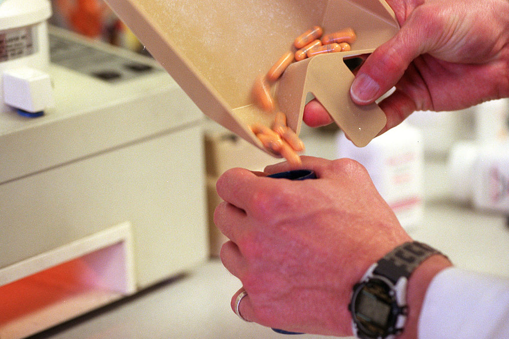 A person&#x27;s hand, wearing a watch, pours orange pills from a tray into a bottle in what appears to be a pharmacy setting