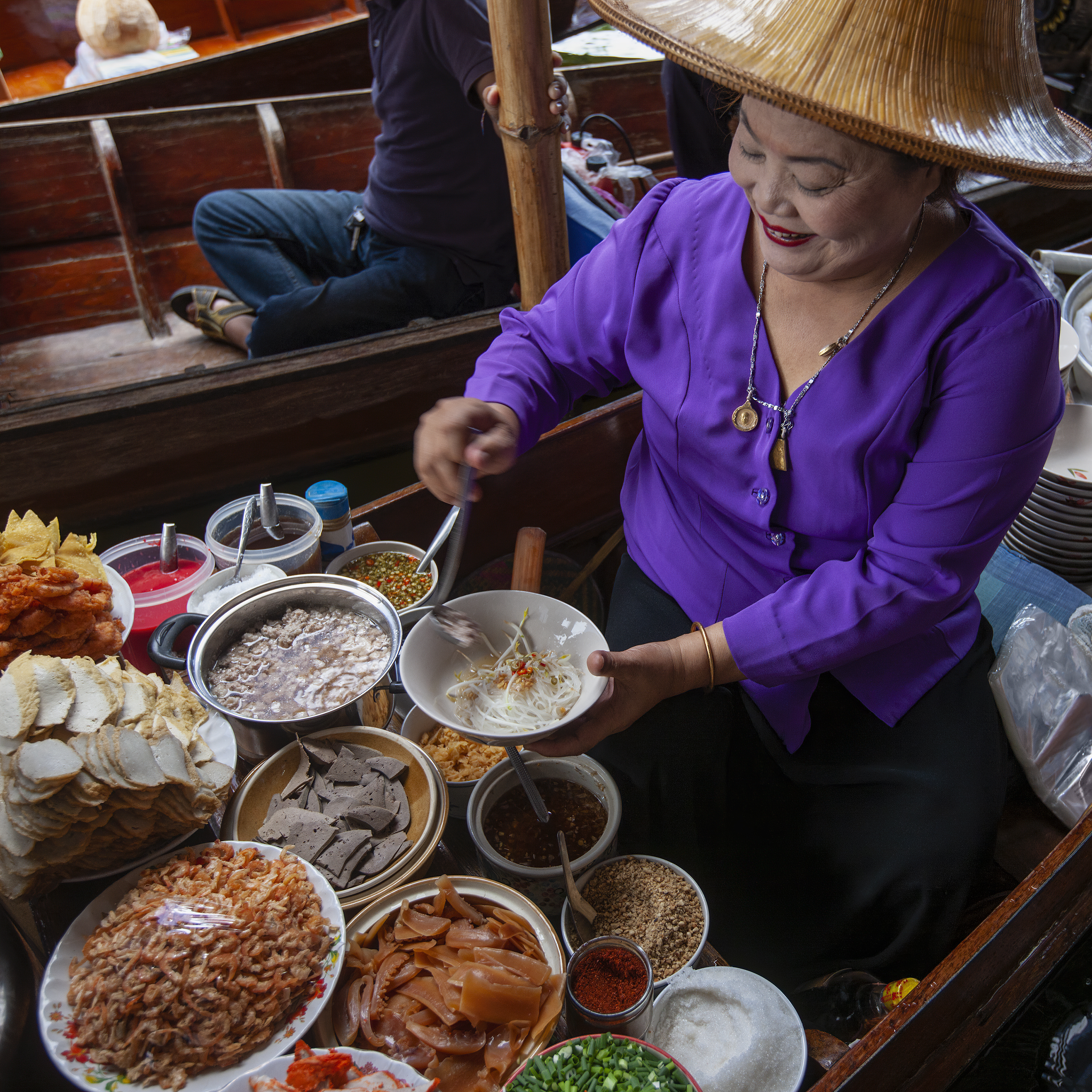 A woman wearing a traditional hat and a purple shirt prepares food at a floating market, surrounded by various colorful ingredients and dishes