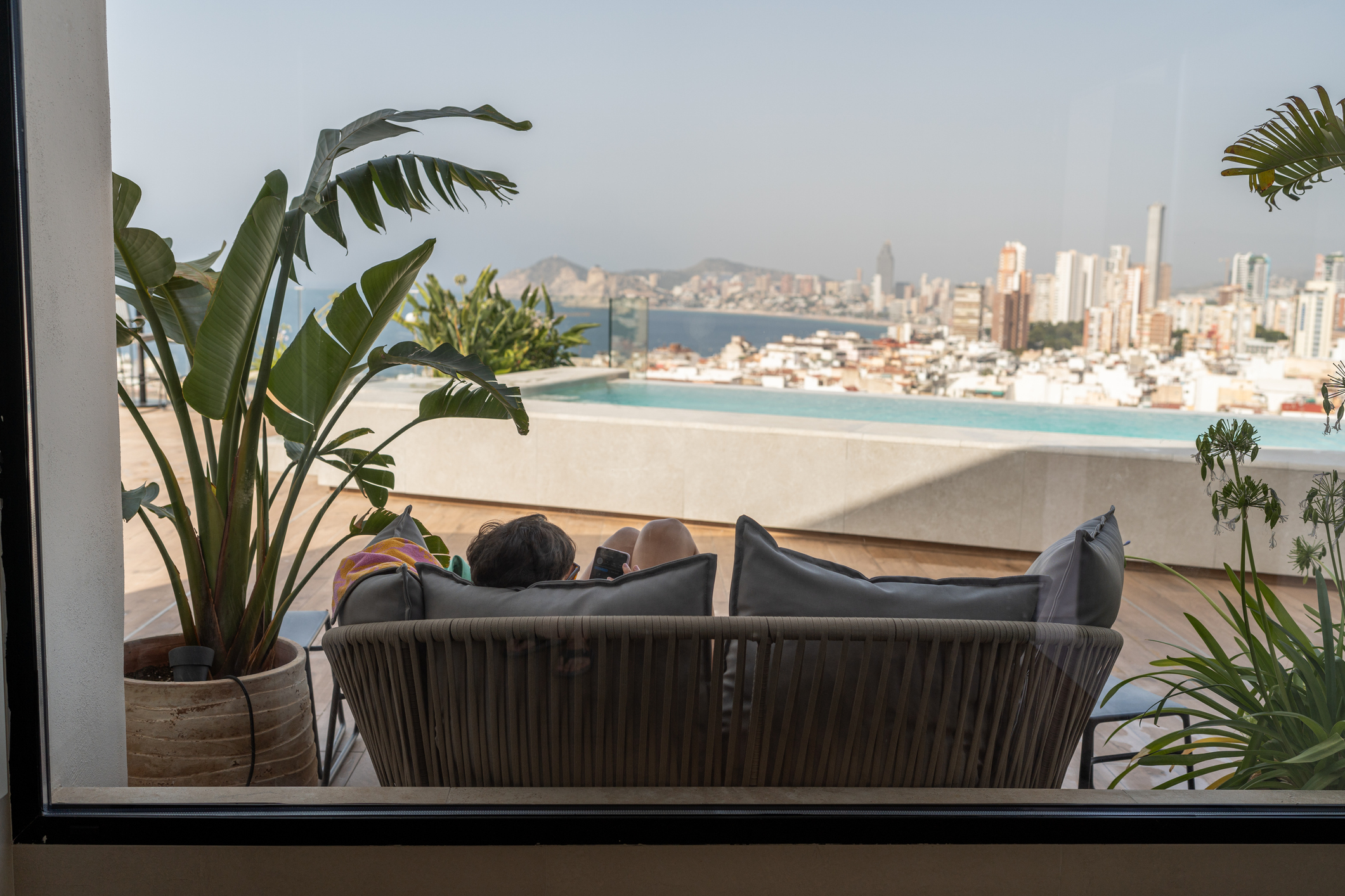 A person is lounging on an outdoor sofa, reading a book beside a rooftop pool, with a city skyline in the background