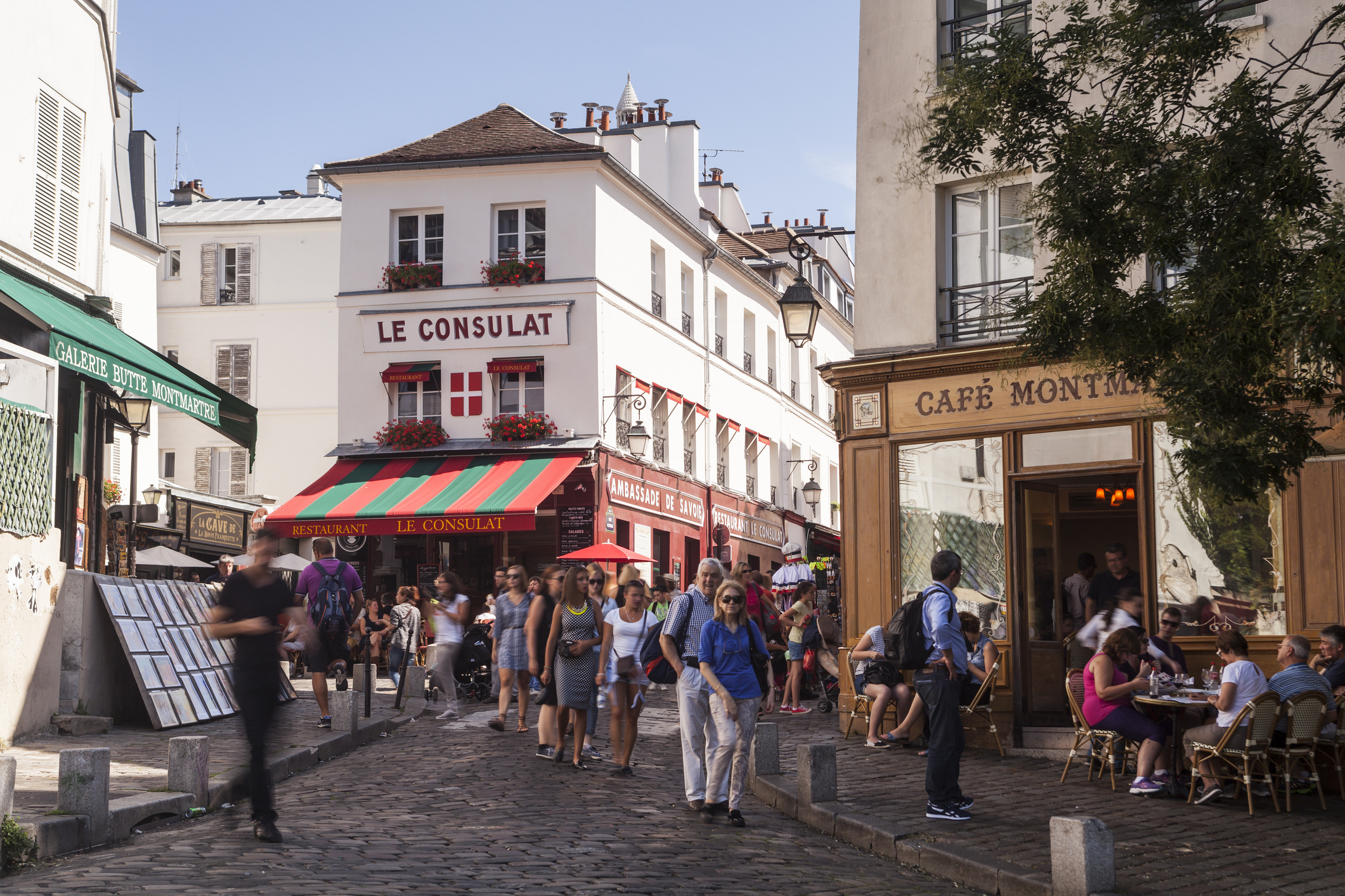 People walking and dining at a busy street in Paris, with cafes and shops, including &quot;Le Consulat&quot; in the background, indicating a typical work and money setting in France