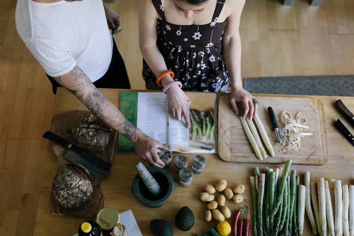 Two people cooking together, referencing a cookbook, surrounded by ingredients including asparagus, potatoes, avocado, and bread on a kitchen counter