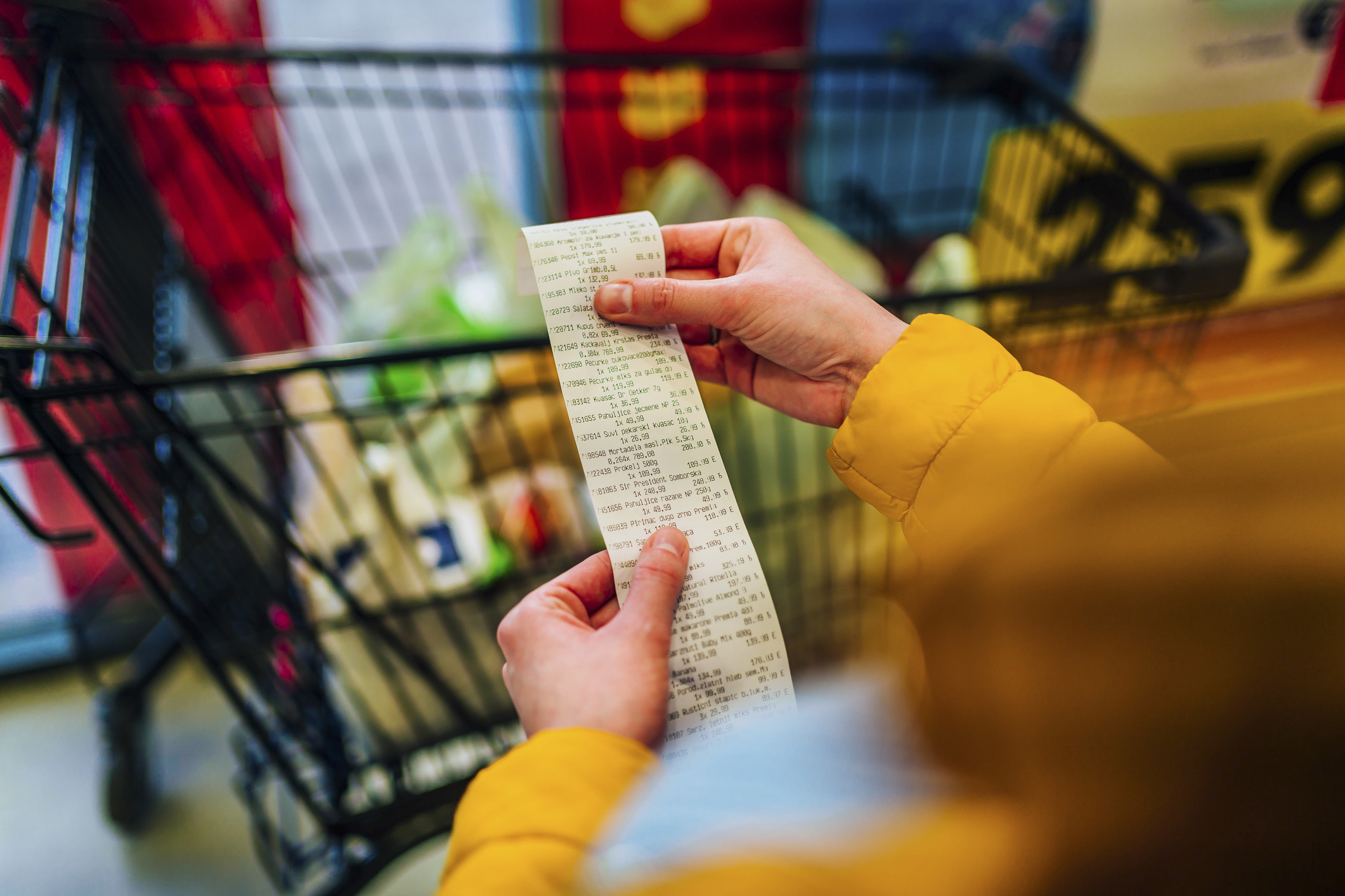 A person holds a receipt and stands in front of a shopping cart filled with groceries