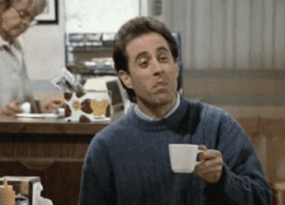 Jerry Seinfeld holds a coffee cup in a diner while making a facial expression