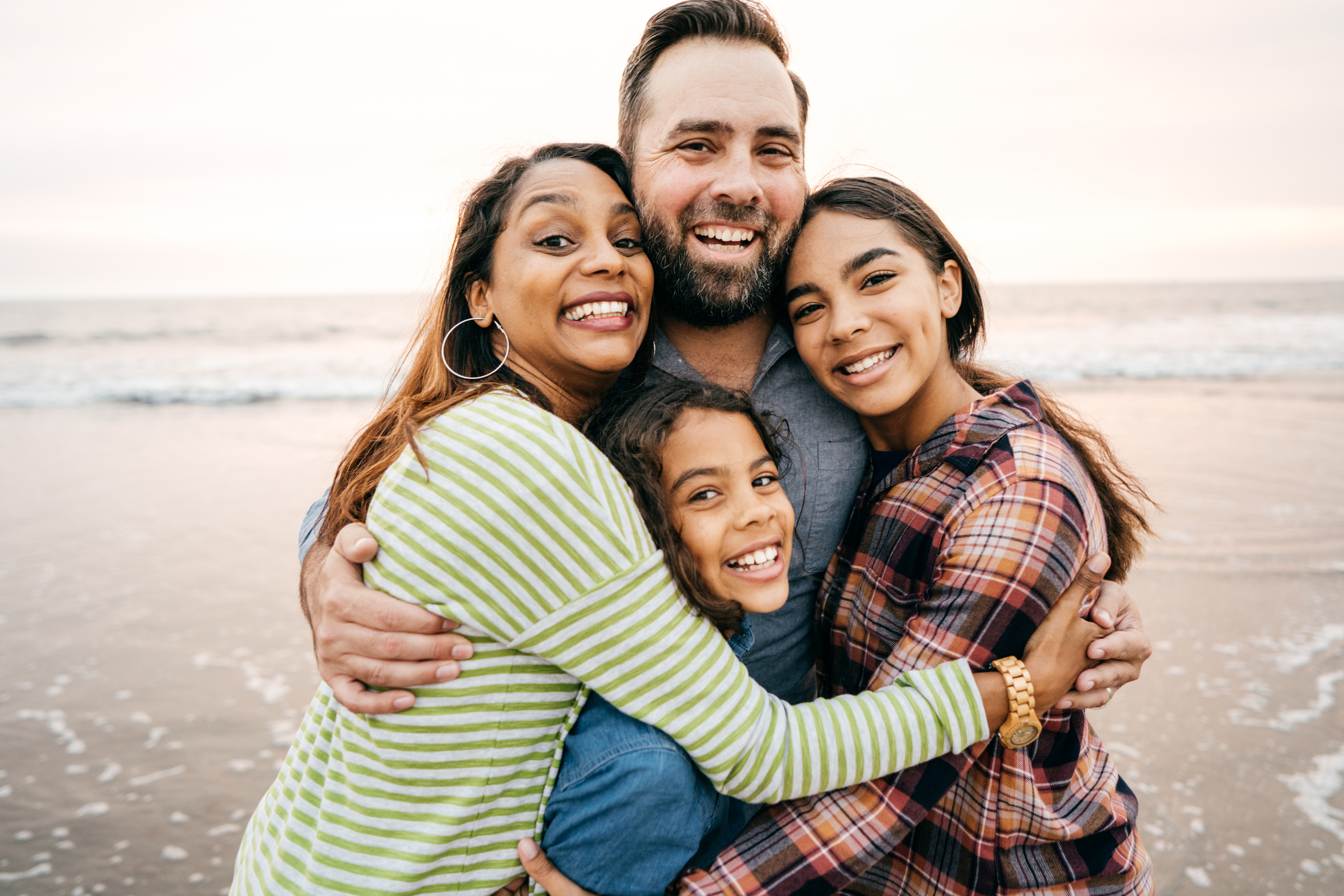 A smiling family of four hugs on a beach, including two children and two adults