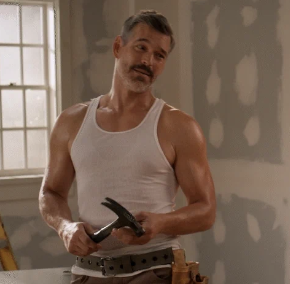Eddie Cibrian wearing a white tank top and brown belt with tools, holds a hammer in a room under construction