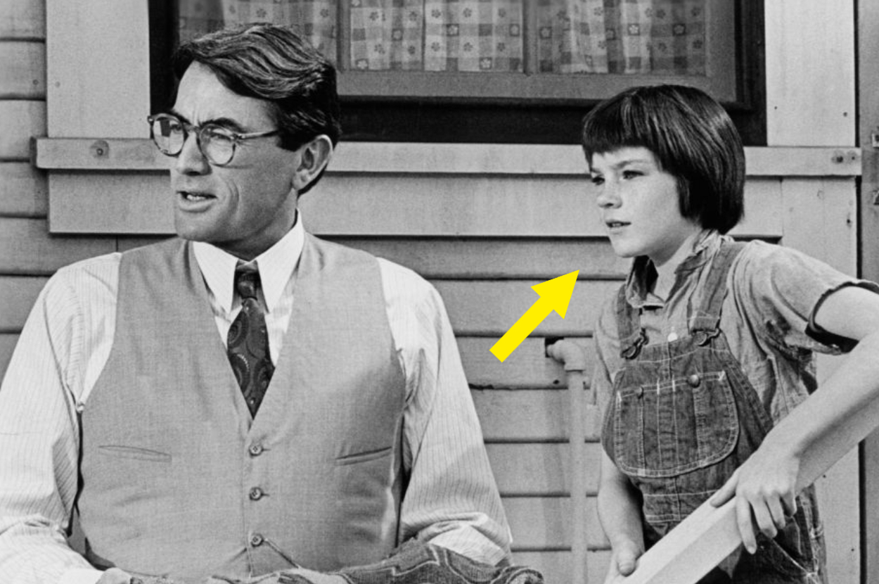 Gregory Peck wearing a vest, tie, and glasses, with Mary Badham in overalls and a short haircut, standing next to him on a porch